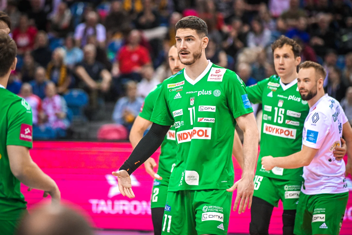 Zawodnicy drużyny siatkarskiej w zielonych strojach z logo sponsorów na froncie przygotowują się do kolejnej akcji na boisku, w tle widoczna jest publiczność kibicująca na trybunach.