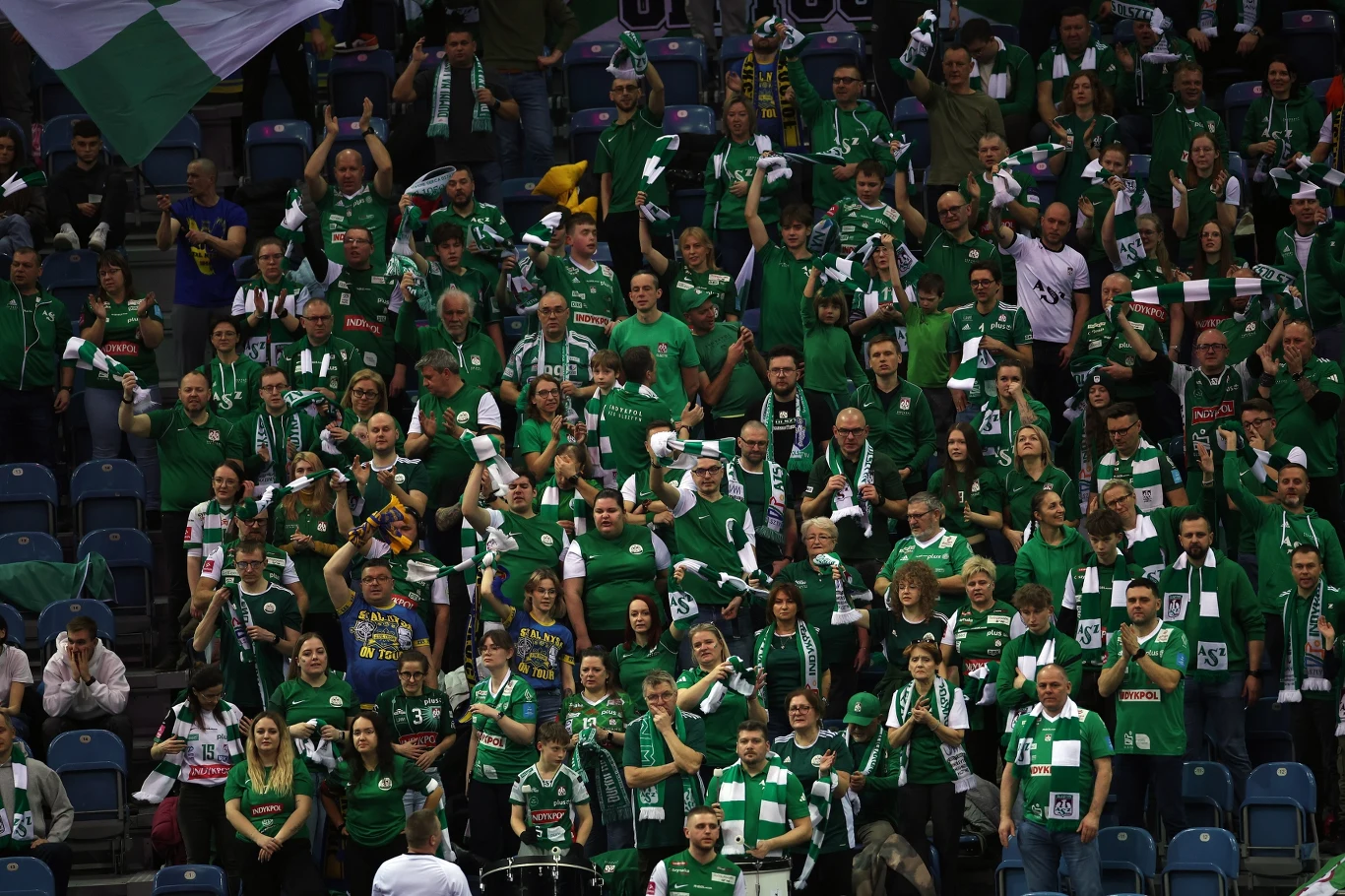 Tłum kibiców ubranych głównie w zielone koszulki i szaliki sportowe, wiwatujących i dopingujących swoją drużynę na trybunach hali sportowej.