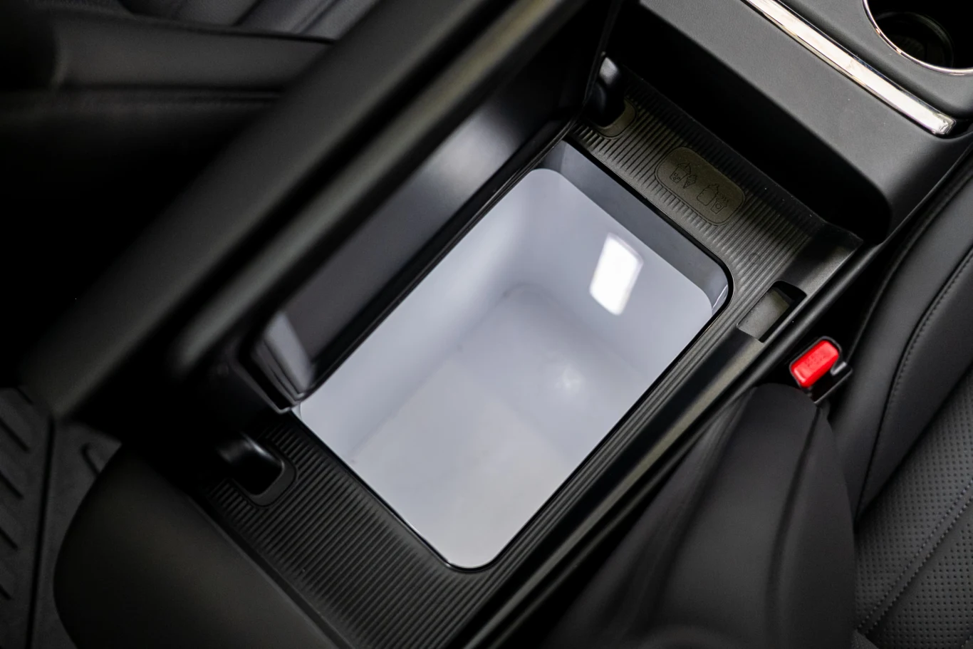 Otwarte wnętrze schowka znajdującego się pomiędzy przednimi fotelami samochodu z widocznym miejscem na przechowywanie przedmiotów. Po bokach znajdują się fotele z czarnej skóry oraz fragment tunelu środkowego z uchwytem na kubek.