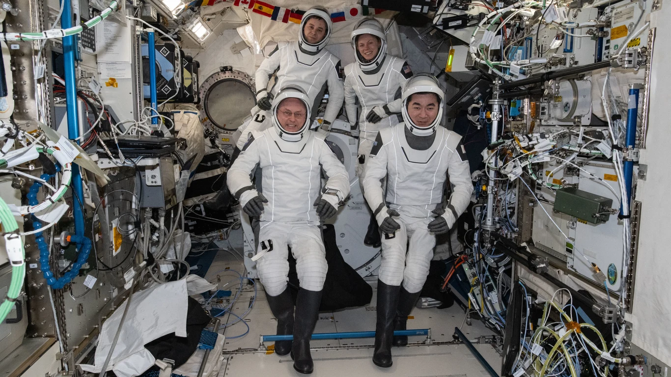 Czterech astronautów w białych skafandrach kosmicznych znajduje się we wnętrzu modułu stacji kosmicznej, otoczeni zaawansowanym sprzętem i instalacjami technicznymi.