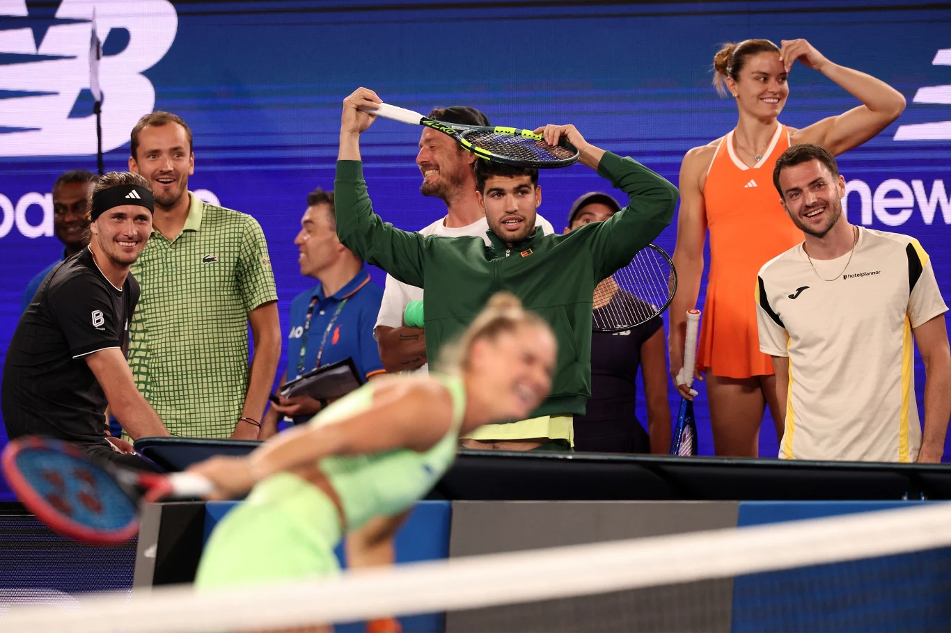 Grupa tenisistów uśmiecha się i żartuje na korcie, w tle widoczne są sportowe stroje i rakiety, a jedna z zawodniczek pochyla się nad siatką do przodu. Panuje swobodna, radosna atmosfera.