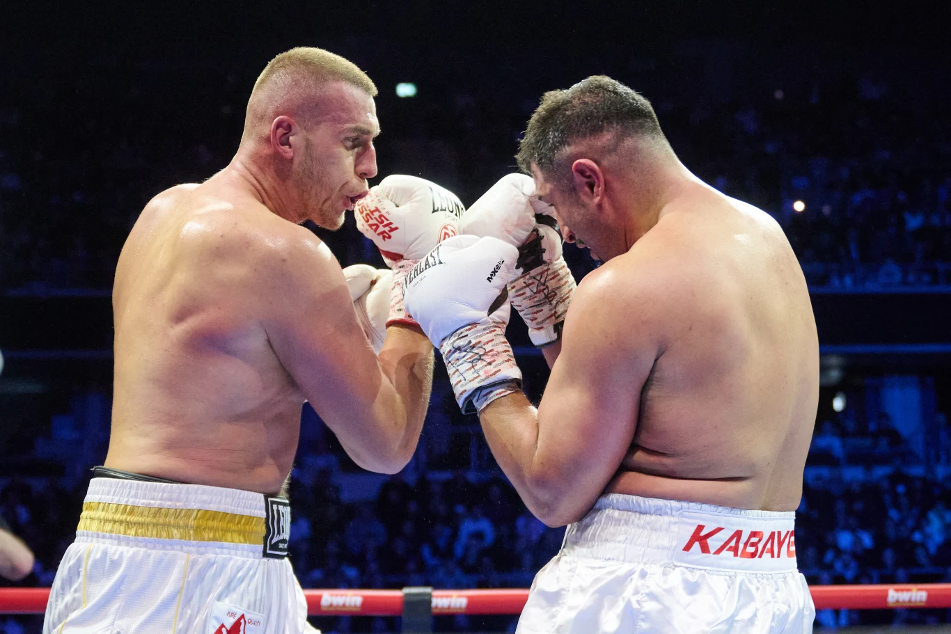 Dwóch bokserów w ringu, obaj w rękawicach i białych spodenkach, skupionych na rywalizacji podczas pojedynku przy sportowym oświetleniu.