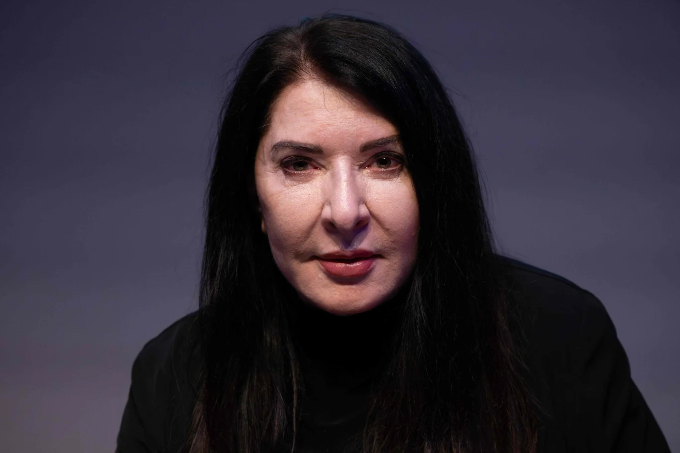 Kobieta o długich, czarnych włosach, ubrana w czarną bluzkę, patrzy prosto w obiektyw na neutralnym, jednolitym tle.