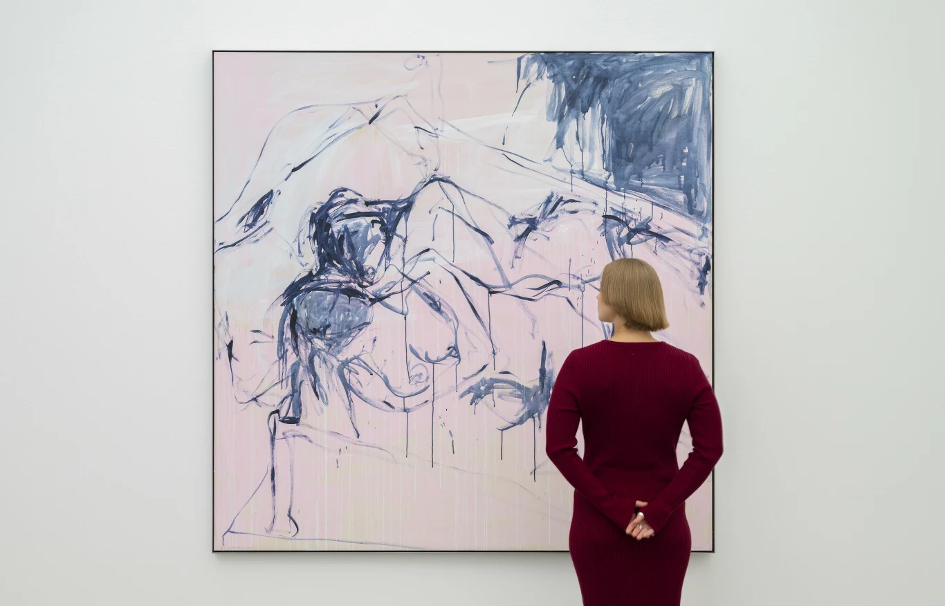 Kobieta w bordowej sukience stoi tyłem do widza i ogląda dużą, abstrakcyjną akwarelę w odcieniach niebieskiego i różu, wiszącą na białej ścianie galerii.