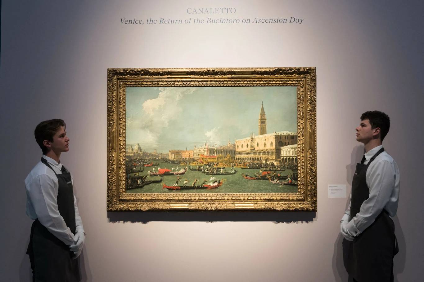 Dzieło sztuki w złotej ramie prezentowane na ścianie galerii, po obu stronach stoją dwaj elegancko ubrani mężczyźni w jednakowych strojach, patrzący na obraz. Na obrazie widoczne jest włoskie miasto z gondolami na wodzie, charakterystyczną architekturą...