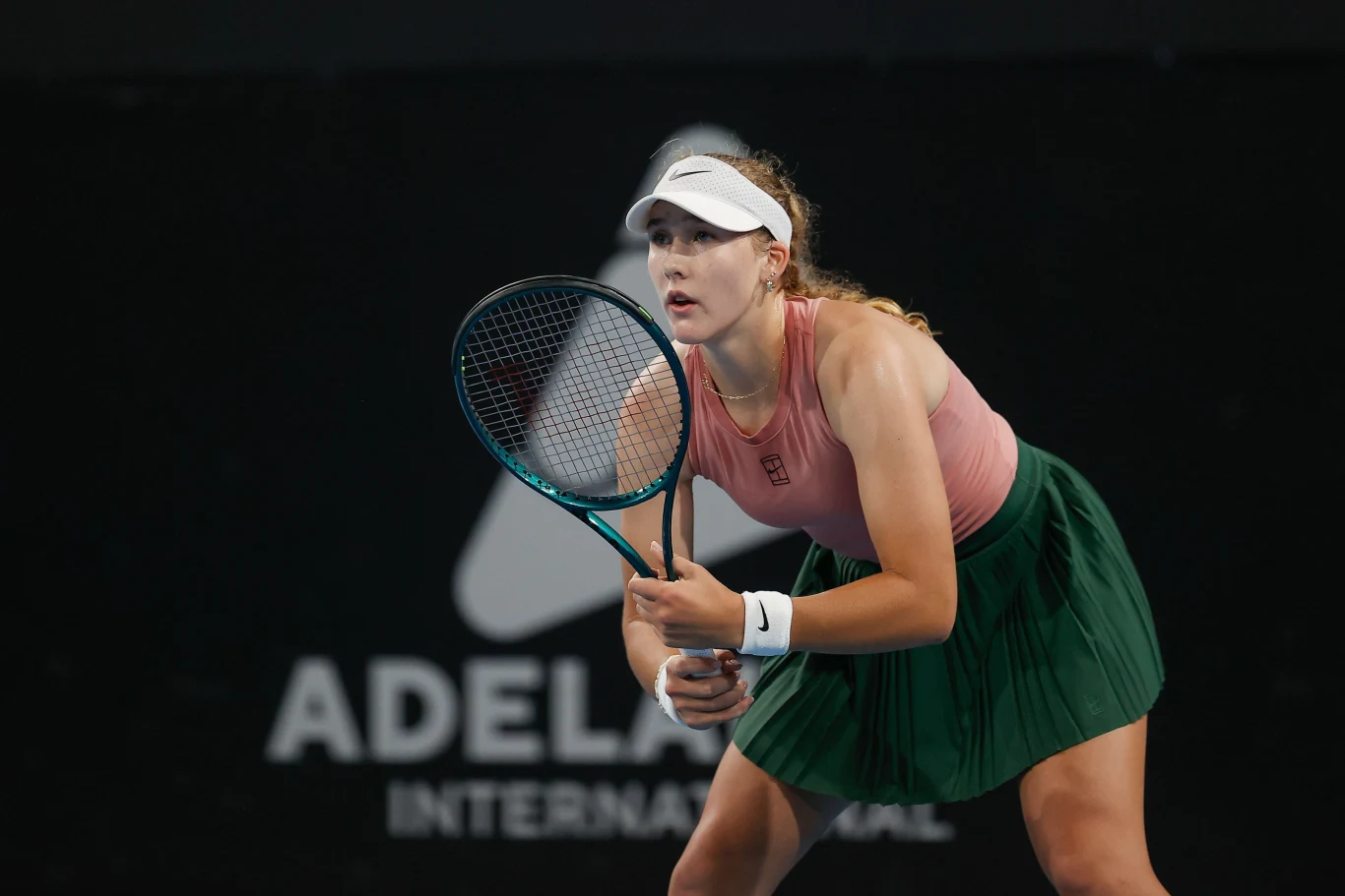 Zawodniczka tenisowa w różowej koszulce i zielonej spódniczce skupiona na korcie, trzyma rakietę w gotowości do odbicia piłki. Na tle widoczne logo turnieju tenisowego Adelaide International.