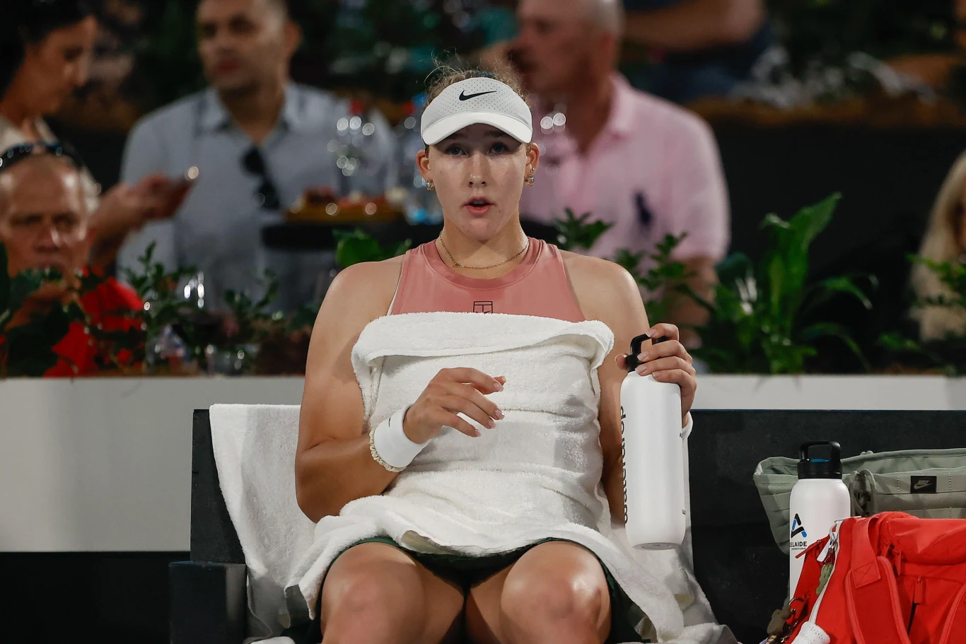 Tenisistka siedzi na ławce podczas przerwy w meczu, trzymając w jednej ręce ręcznik, a w drugiej butelkę z wodą. Ubrana w sportowy strój oraz czapkę z daszkiem. W tle widoczne zamazane postacie kibiców.