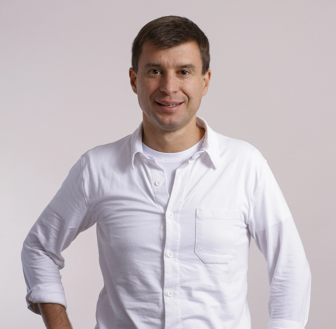 Mężczyzna w białej koszuli, stojący na tle jasnego, jednolitego tła, lekko uśmiechnięty, z jedną ręką opartą na biodrze.