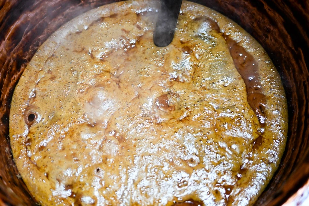 Gotująca się gęsta masa o brązowej barwie w dużym garnku, na powierzchni widoczne są bąble i parująca piana, co sugeruje proces przygotowywania np. karmelu, syropu lub innej gęstej substancji.