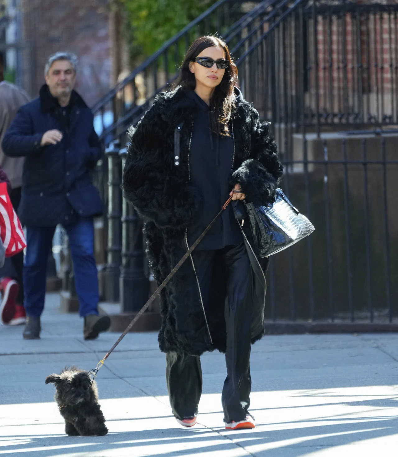 Elegancka kobieta w długim, czarnym futrze prowadzi na smyczy małego czarnego psa, spacerując po miejskim chodniku. W ręku trzyma stylową torebkę, a na twarzy ma ciemne okulary przeciwsłoneczne.