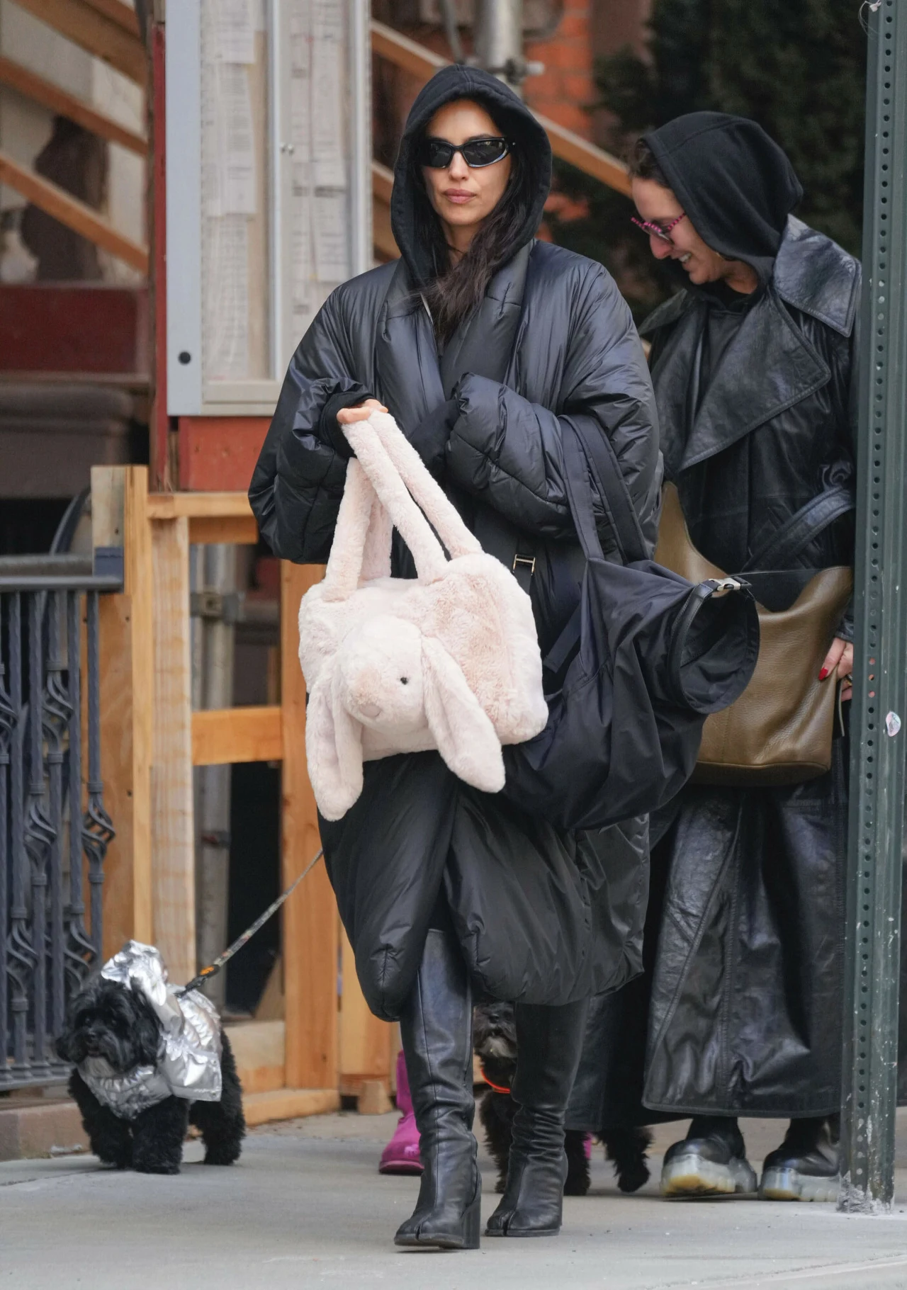 Kobieta w długim, czarnym, puchowym płaszczu i wysokich butach trzyma miękką torbę w kształcie królika. Ma na sobie ciemne okulary przeciwsłoneczne i idzie chodnikiem w towarzystwie osoby w czarnym płaszczu. Obok nich pies ubrany w srebrny kubrak prowa...