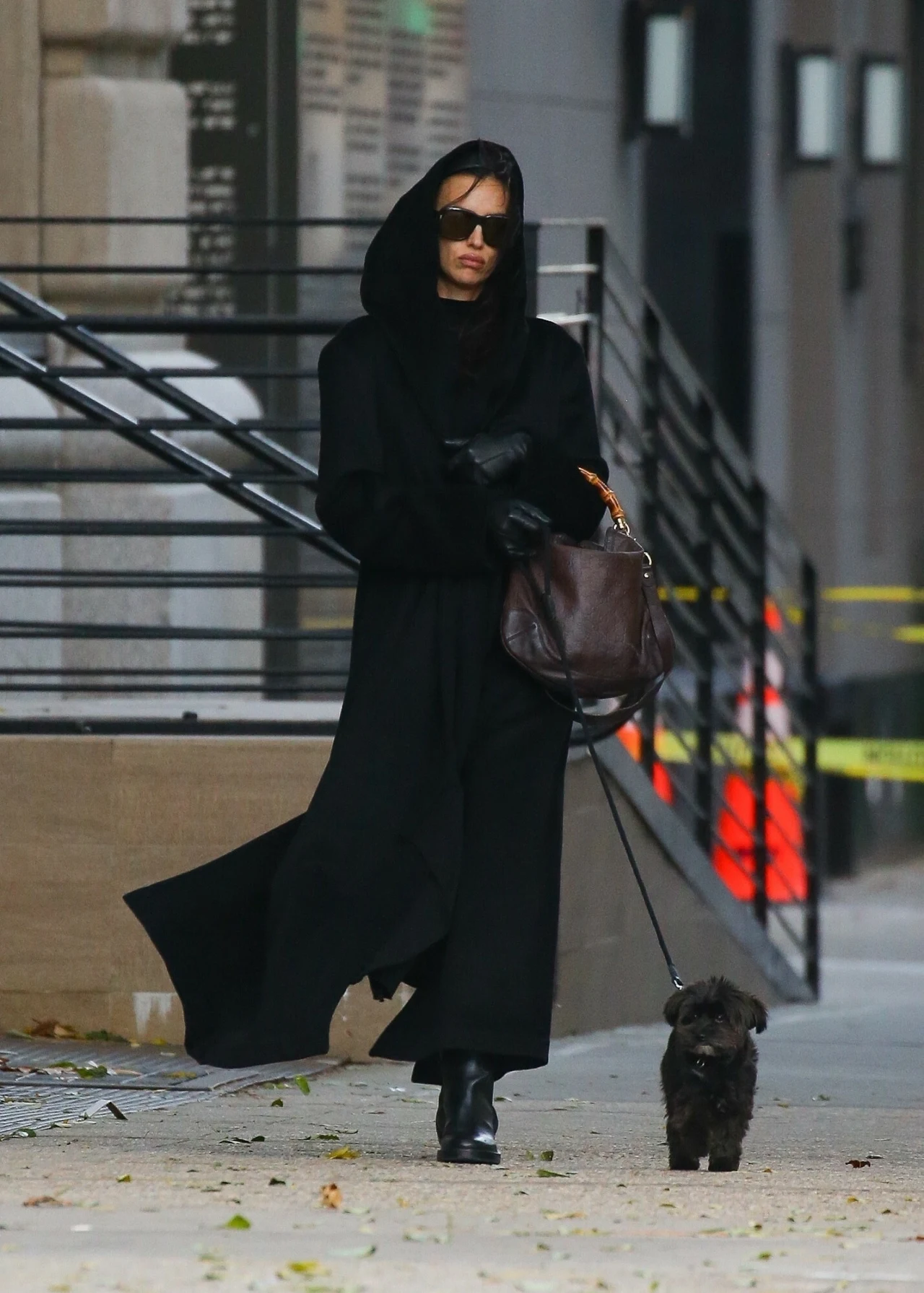Kobieta ubrana w długi, czarny płaszcz z kapturem i ciemne okulary spaceruje z małym czarnym psem po miejskim chodniku. W tle znajdują się schody, poręcze oraz żółta taśma ostrzegawcza.