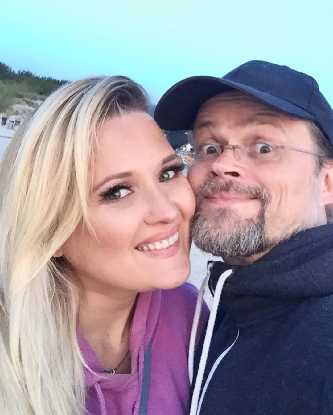 Para uśmiechniętych osób robi selfie na tle jasnego nieba i pagórkowatego terenu z roślinnością, oboje ubrani w sportowe bluzy, mężczyzna ma czapkę z daszkiem i okulary.