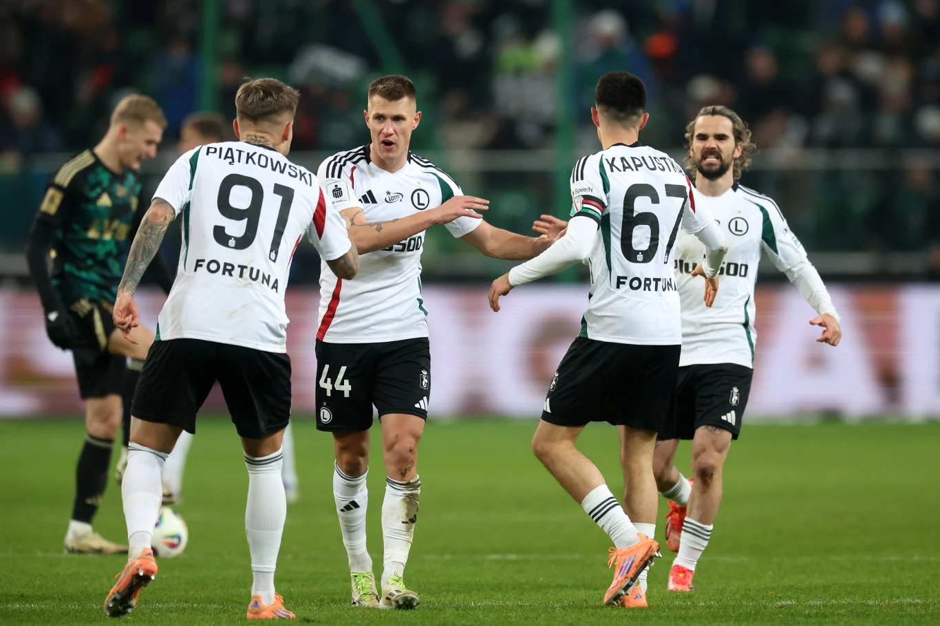 Piłkarze w białych strojach z zielonymi i czerwonymi akcentami świętują wspólnie akcję podczas meczu piłkarskiego, wyrażając radość oraz sportową rywalizację. W tle widoczni są kibice oraz inni zawodnicy.