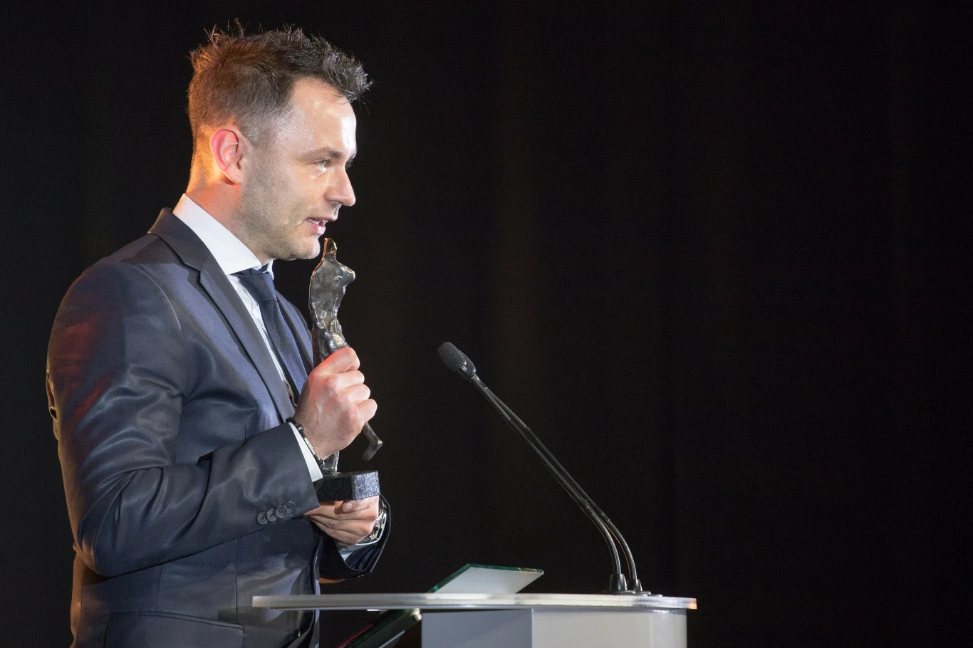 Igor Klaja, 13.01.2018 r. Mężczyzna w garniturze stoi przy mównicy, trzyma statuetkę w jednej ręce i mówi do mikrofonu, wyrażając emocje związane z odbiorem nagrody. Ciemne tło skupia uwagę na jego sylwetce.