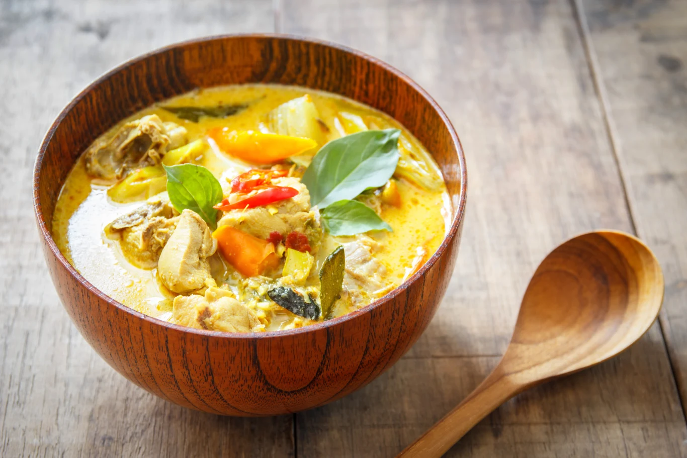 Drewniana miska wypełniona żółtym curry z kawałkami kurczaka, warzyw i świeżych liści bazylii, obok znajduje się drewniana łyżka leżąca na drewnianym stole.