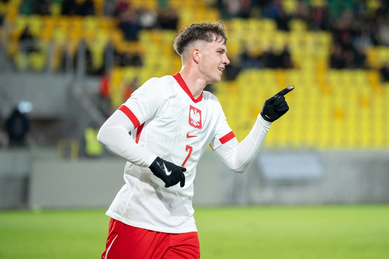 Młody piłkarz w białym stroju z czerwoną lamówką i numerem 7 świętuje zdobycie bramki, wskazując palcem przed siebie, w tle rozmyte żółte trybuny stadionu.