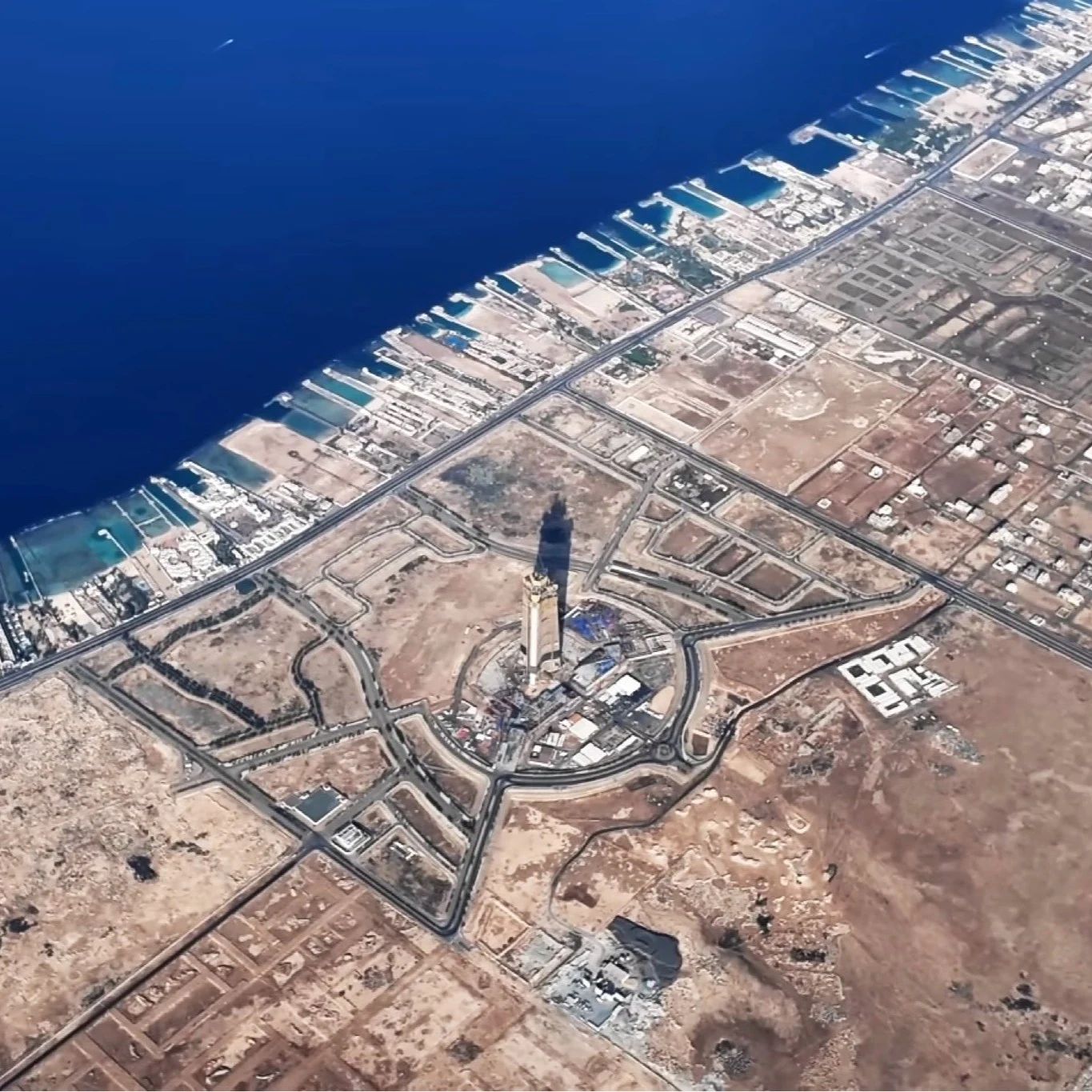 Jeddah Tower z góry wygląda już bardzo imponująco. Ogromny wieżowiec cały czas rośnie. Widok z lotu ptaka na pustynne tereny, nowoczesną zabudowę oraz pojedynczy bardzo wysoki wieżowiec otoczony siecią dróg. W tle wybrzeże morza z licznymi marinami i turkusowym kolorem wody.