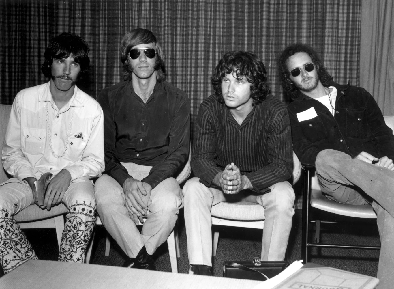 The Doors w 1968 r. - od lewej: John Densmore, Ray Manzarek, Jim Morrison i Robby Krieger Czterech mężczyzn siedzi na krzesłach w pomieszczeniu, trzech z nich nosi okulary przeciwsłoneczne, wszyscy są w luźnych ubraniach, jeden trzyma zeszyt, a za nimi znajdują się zasłony.