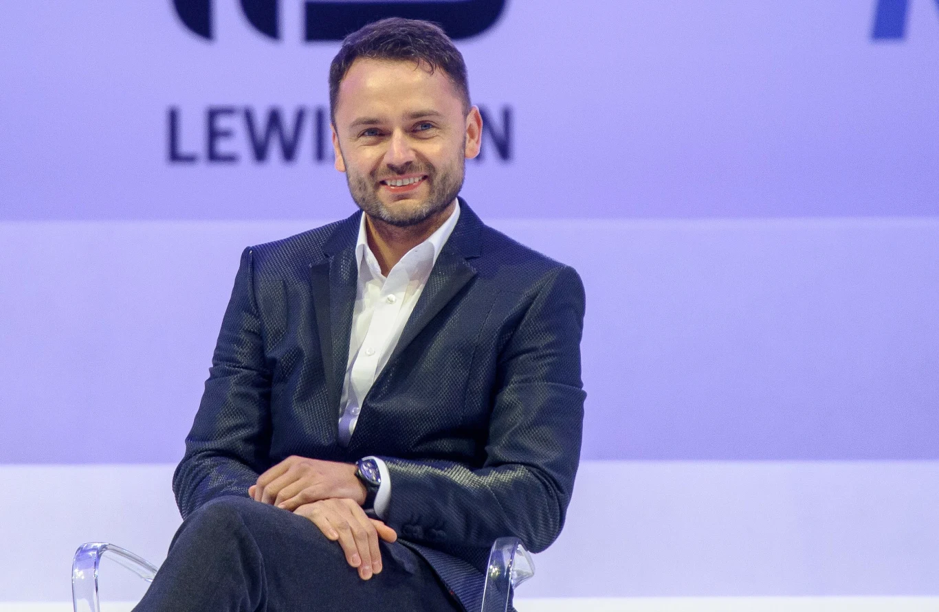 Igor Klaja, 15.06.2016 r. Mężczyzna w eleganckim garniturze siedzący na przezroczystym krześle, uśmiechnięty, w tle jasna plansza z częściowo widocznymi napisami i logotypami.