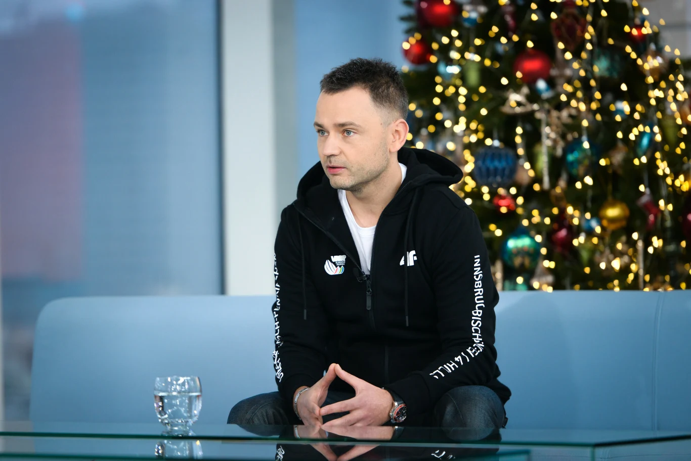 Igor Klaja, 16.12.2018 r. Mężczyzna siedzący na jasnoniebieskiej sofie w studiu telewizyjnym, ubrany w czarną bluzę z zamkiem, w tle udekorowana choinka z kolorowymi bombkami i światełkami, na stoliku przezroczysta szklanka z wodą.