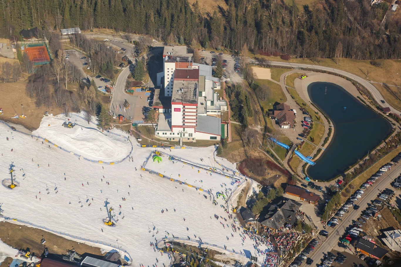 Stok narciarski z licznymi osobami uprawiającymi narciarstwo, położony obok dużego hotelu i sztucznego zbiornika wodnego, wokół którego znajdują się trasy spacerowe oraz kilka budynków i parking. W tle widoczne lasy.
