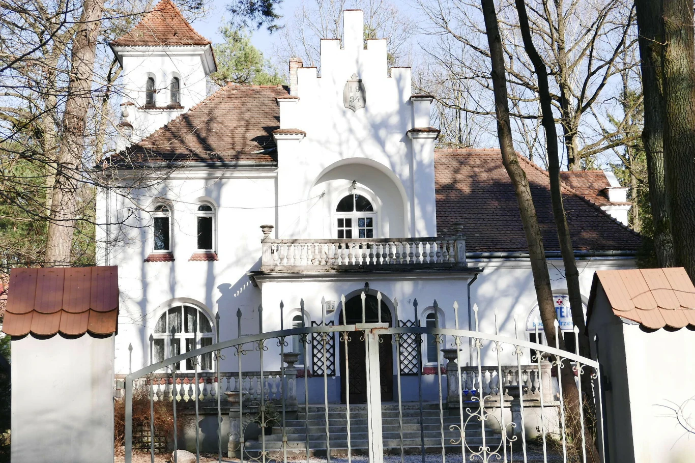 Biała willa z zabytkowymi elementami architektonicznymi, otoczona metalowym ogrodzeniem, położona wśród drzew, z widocznym balkonem i tarasem, dach kryty czerwoną dachówką.