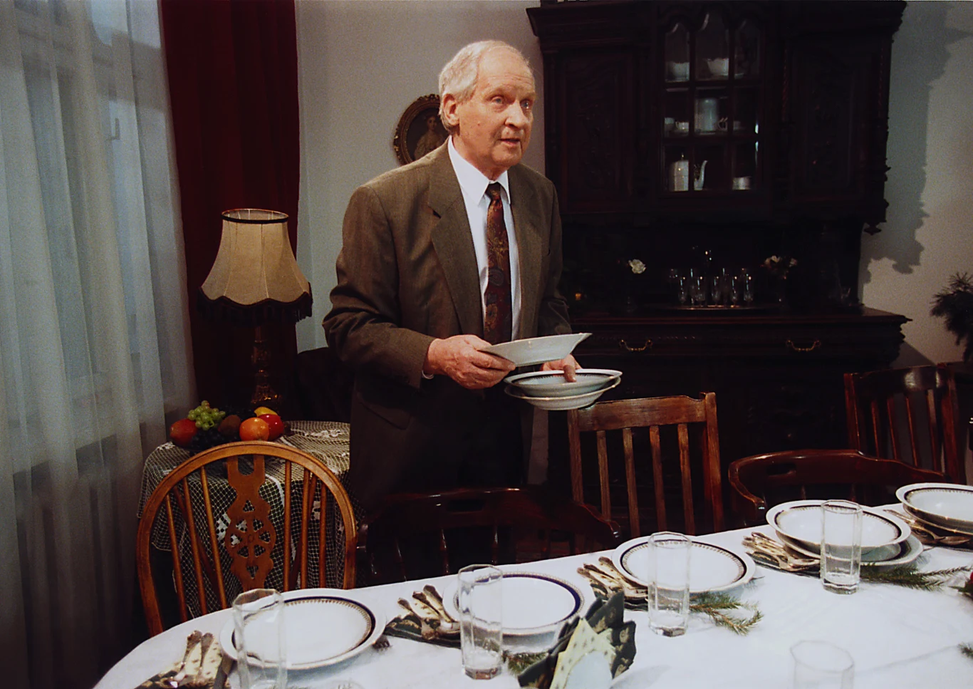 Starszy mężczyzna w garniturze ustawia talerze na uroczystym stole w jadalni. Na stole leżą talerze, szklanki, sztućce i dekoracje, a w tle stoi kredens z zastawą. Całość tworzy atmosferę przygotowań do ważnego posiłku lub rodzinnej uroczystości.