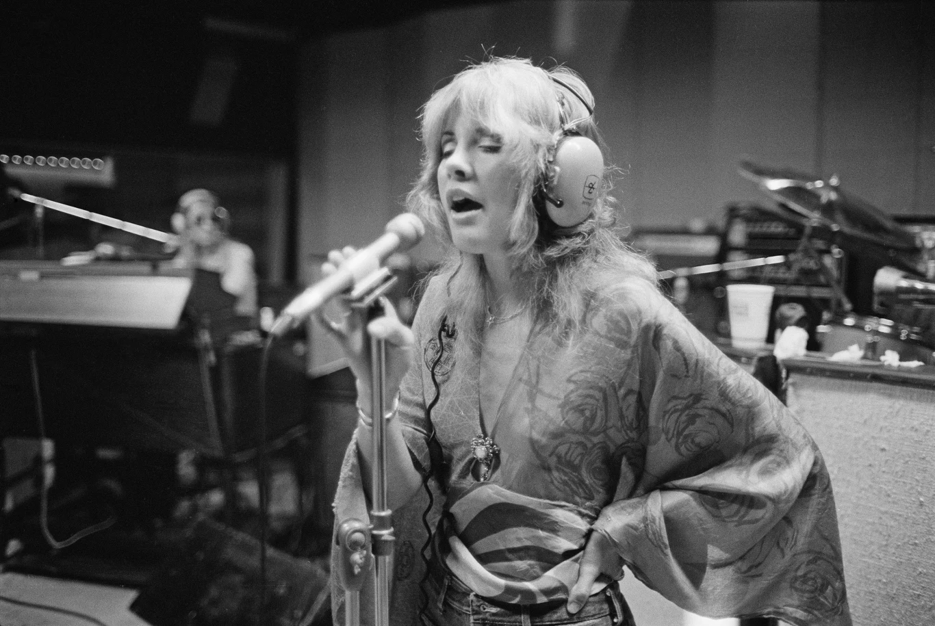 Stevie Nicks (Fleetwood Mac) w 1975 r. Kobieta w słuchawkach śpiewa do mikrofonu w studiu nagraniowym, w tle widoczny muzyk grający na klawiaturze oraz instrumenty muzyczne.