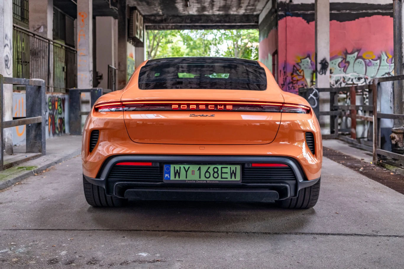Pomarańczowy samochód Porsche Taycan Turbo S zaparkowany w industrialnym, opuszczonym budynku z widocznymi ścianami pokrytymi kolorowym graffiti, tylne światła auta wyróżniają się na tle betonowej przestrzeni.