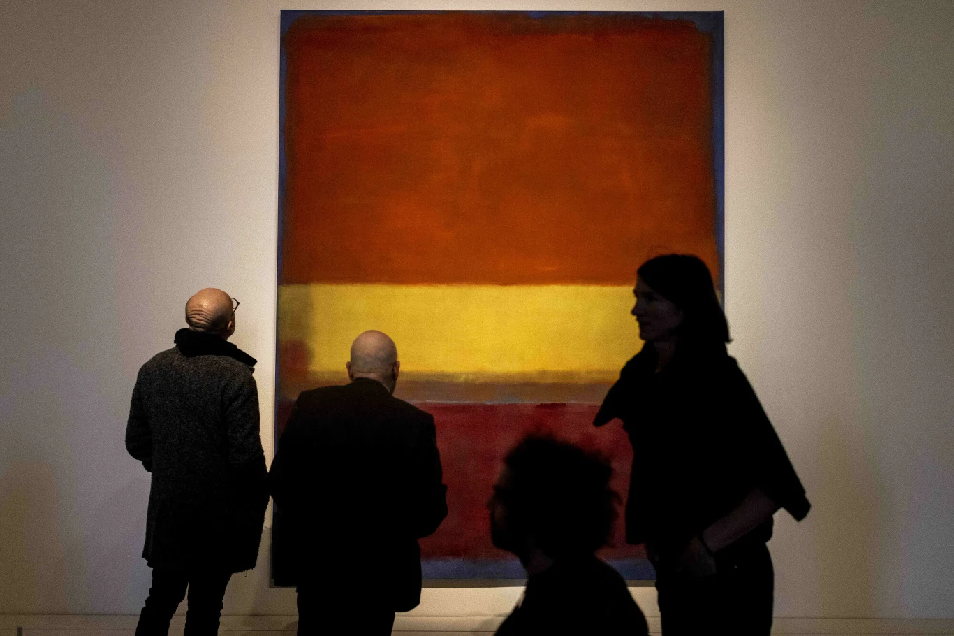 Grupa osób podziwia duży abstrakcyjny obraz o intensywnych, ciepłych barwach, przedstawiający geometryczne bloki kolorów w odcieniach czerwieni, żółci i pomarańczy na tle ściany galerii.