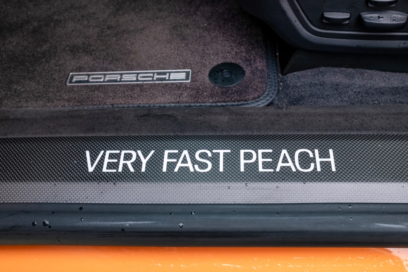 Wnętrze samochodu marki Porsche z widocznym napisem 'VERY FAST PEACH' na progu drzwi oraz dywanikiem z logo producenta, wykończone w ciemnym kolorze z akcentami pomarańczowymi.