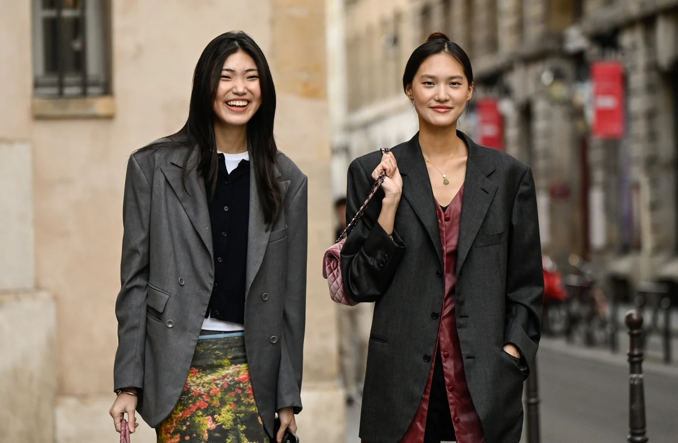 Jak nosić marynarkę w 2026 roku? Dwie młode kobiety ubrane w modne, oversize'owe marynarki, stoją obok siebie na ulicy miejskiej, obie uśmiechnięte, jedna ma na sobie wzorzystą spódnicę, druga trzyma różową torebkę na ramieniu.