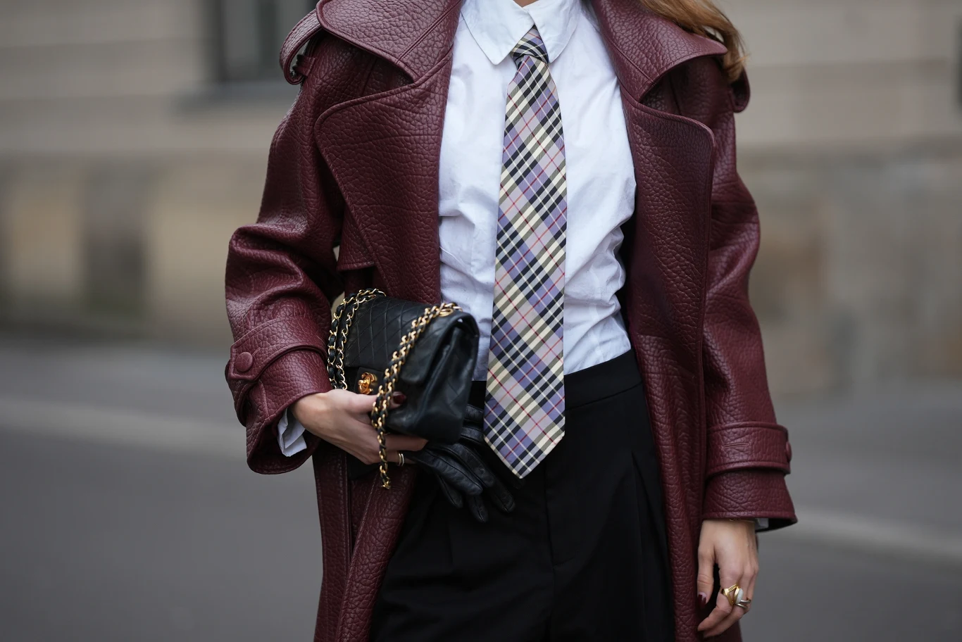 Jak stworzyć modną stylizację z białą koszulą? Elegancka kobieta ubrana w bordowy płaszcz z wyrazistą fakturą, białą koszulę i krawat w kratę, trzyma czarną torebkę na łańcuszku oraz nosi czarne rękawiczki. Stylizacja wyróżnia się nowoczesnym podejściem do klasycznej mody.
