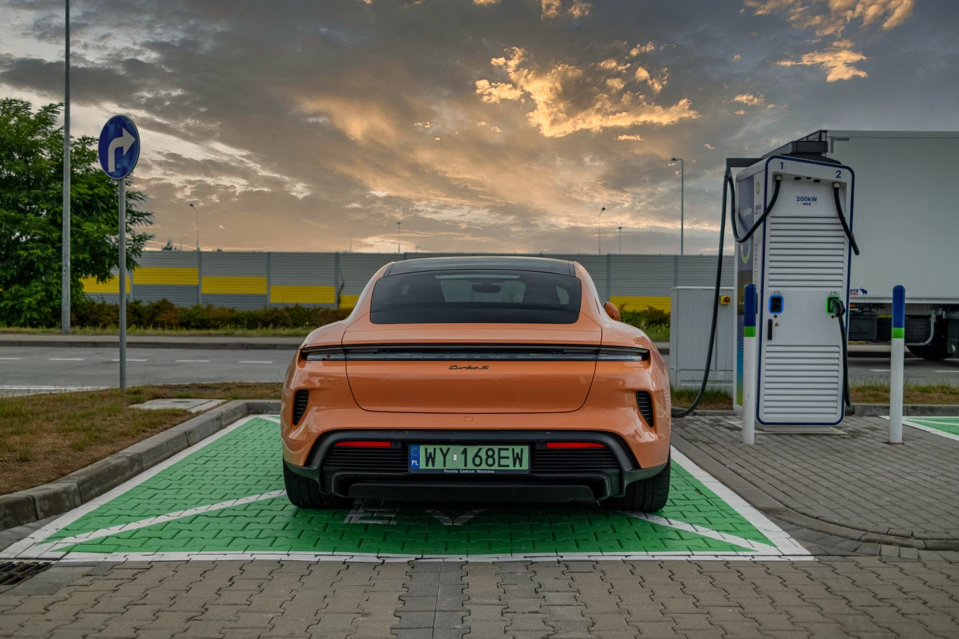 Pomarańczowy samochód elektryczny marki Porsche stoi zaparkowany na miejscu przeznaczonym do ładowania elektrycznych pojazdów. Auto jest podłączone do stacji ładowania, a w tle widoczny jest fragment parkingu, ciężarówka i niebo z chmurami o zachodzie ...