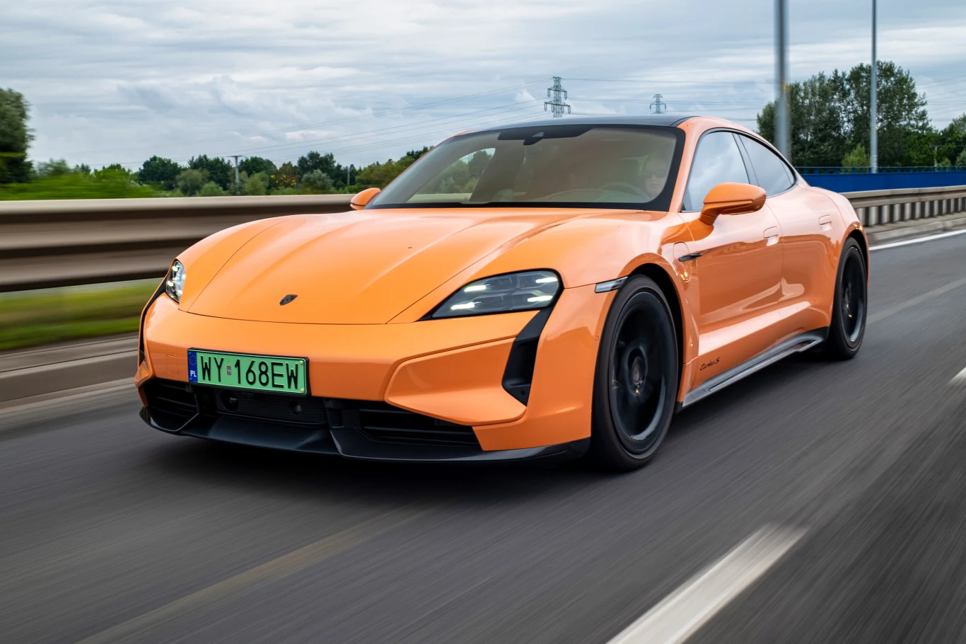 Pomarańczowy samochód sportowy poruszający się po drodze szybkiego ruchu, nowoczesny design, czarne felgi, tło to rozmyte drzewa i barierki, tablica rejestracyjna z zielonym elementem.