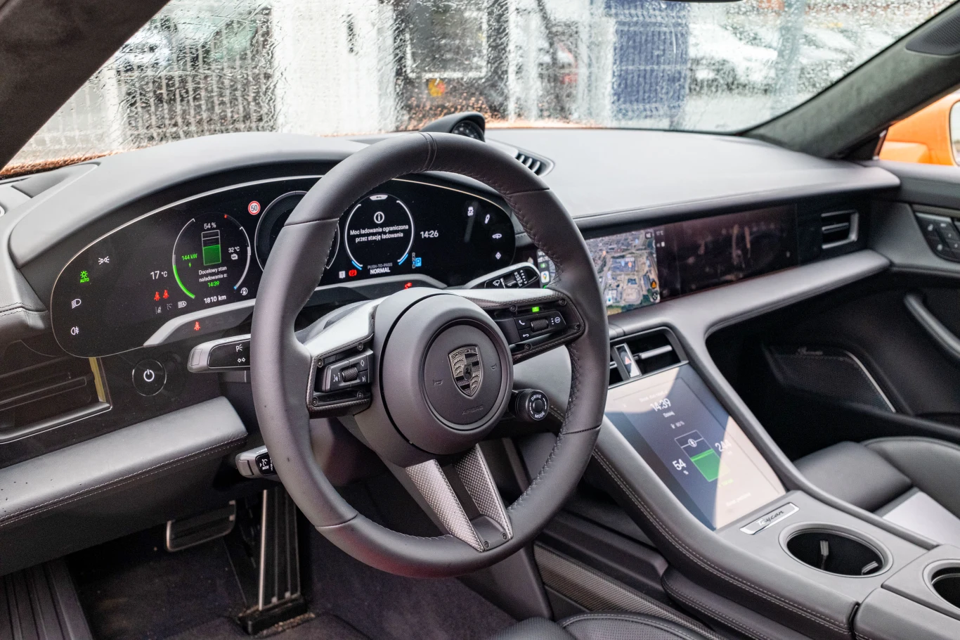 Luksusowe wnętrze samochodu sportowego z elektronicznym, nowoczesnym kokpitem, dużym dotykowym wyświetlaczem centralnym oraz kierownicą z logo producenta. Na panelu widoczne cyfrowe zegary, centralny ekran nawigacji i kontrolery na konsoli środkowej.