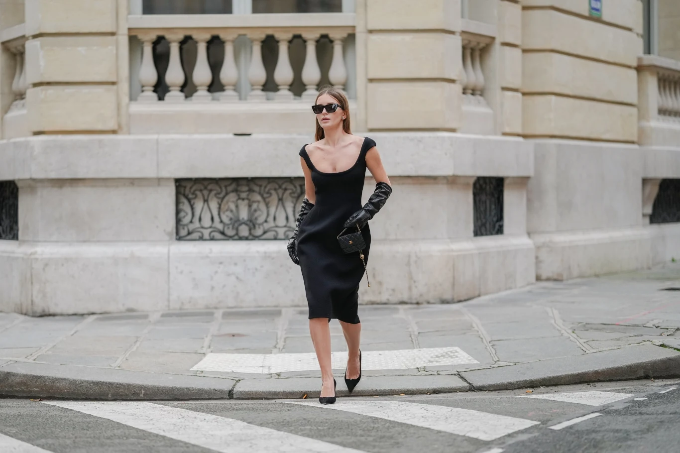 Mała czarna to klasyk, który nigdy się nie znudzi Stylowa kobieta w czarnej sukience i okularach przeciwsłonecznych idzie chodnikiem w eleganckim, miejskim otoczeniu. Trzyma czarną torebkę i nosi czarne rękawiczki, tło stanowi jasny budynek z kamiennymi balustradami.