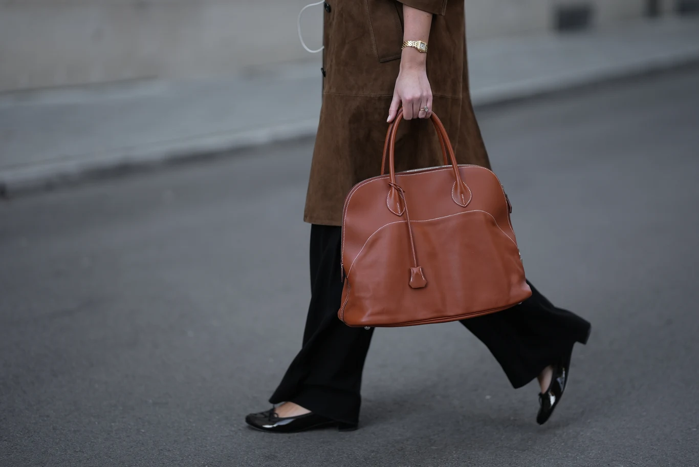 Najmodniejsze torby do pracy Kobieta idąca po ulicy z brązową torebką w dłoni, ubrana w brązowy płaszcz, czarne spodnie i czarne lakierowane buty na płaskim obcasie.