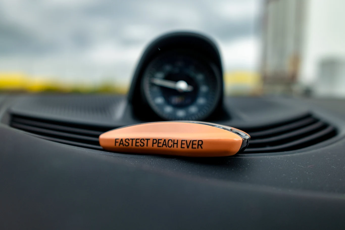 Pomarańczowy przedmiot z napisem FASTEST PEACH EVER leżący na czarnej desce rozdzielczej samochodu, w tle widoczny okrągły zegar umieszczony centralnie.