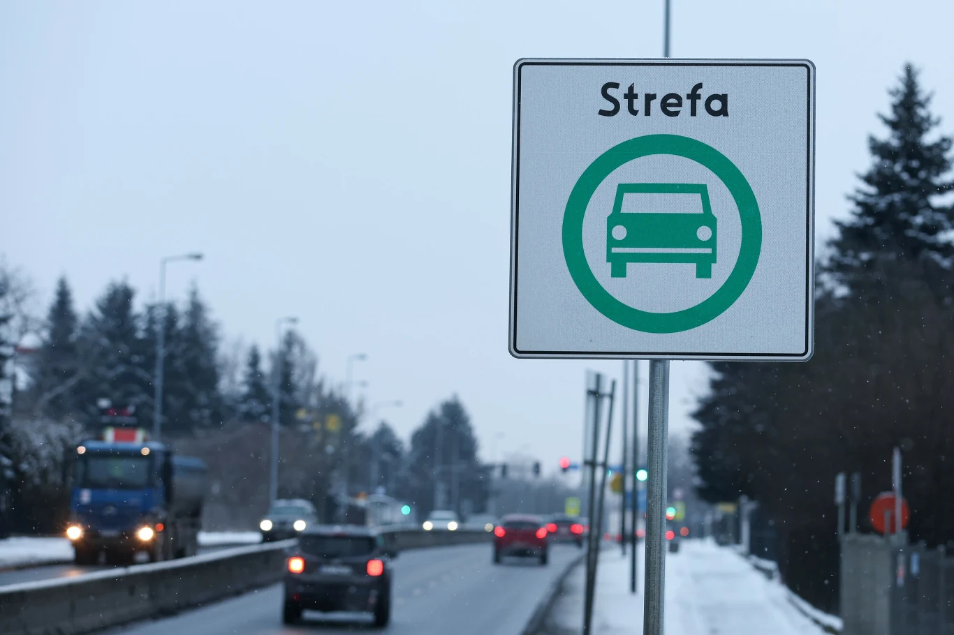 Strefa Czystego Transportu w Krakowie działa od 1.01.2026 r. i jest zgodna z prawem - orzekł sąd Znak drogowy informujący o strefie, na tle ulicy z kilkoma jadącymi samochodami, w zimowej scenerii z widocznymi drzewami i lekko zaśnieżonym poboczem.