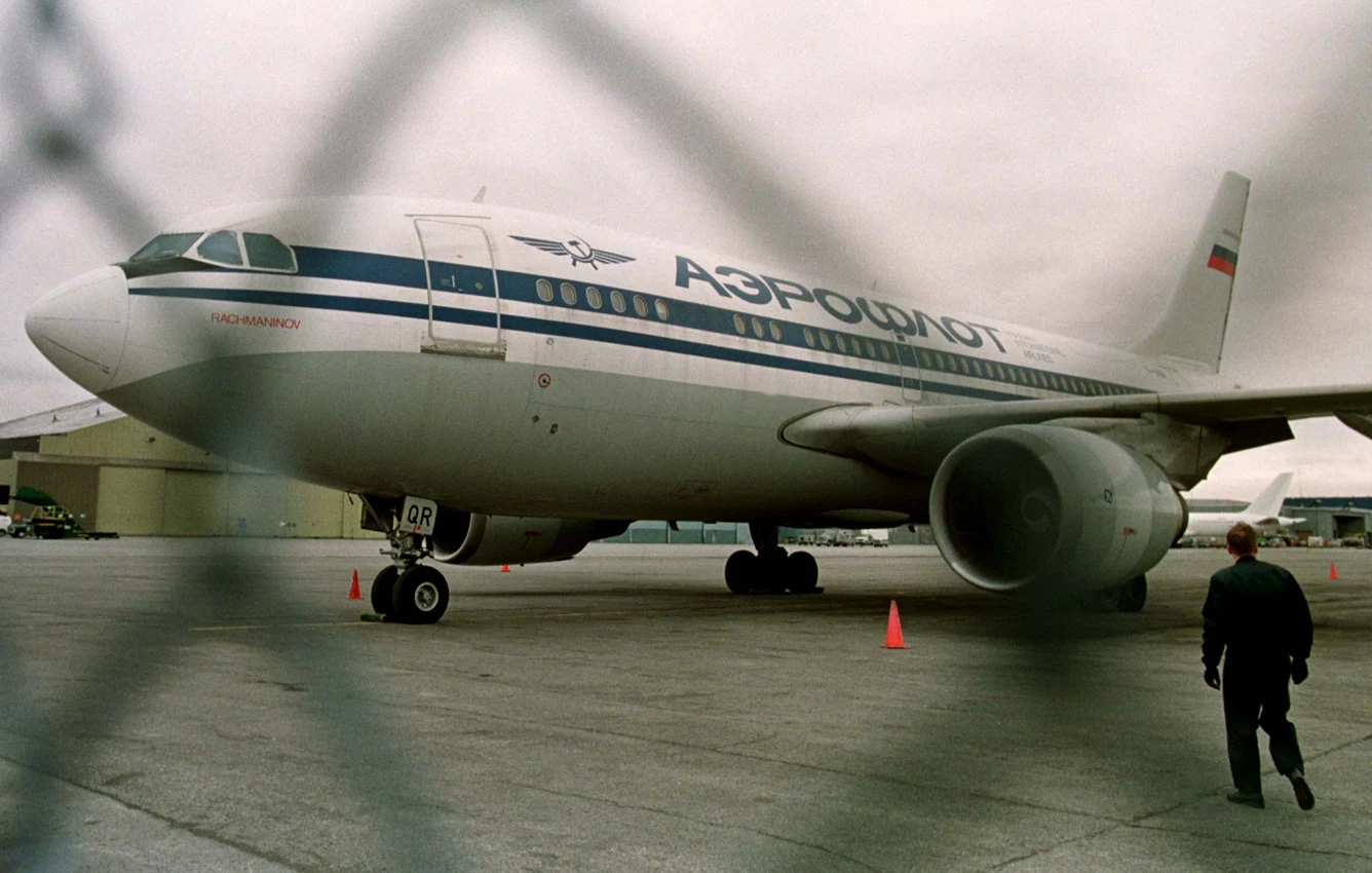 Samolot pasażerski linii Aerofłot stoi na płycie lotniska, otoczony pachołkami bezpieczeństwa. Wchodząca z prawej strony osoba zbliża się do maszyny. W tle widoczne hangary lotniskowe, a całość obserwowana przez siatkę ogrodzenia.