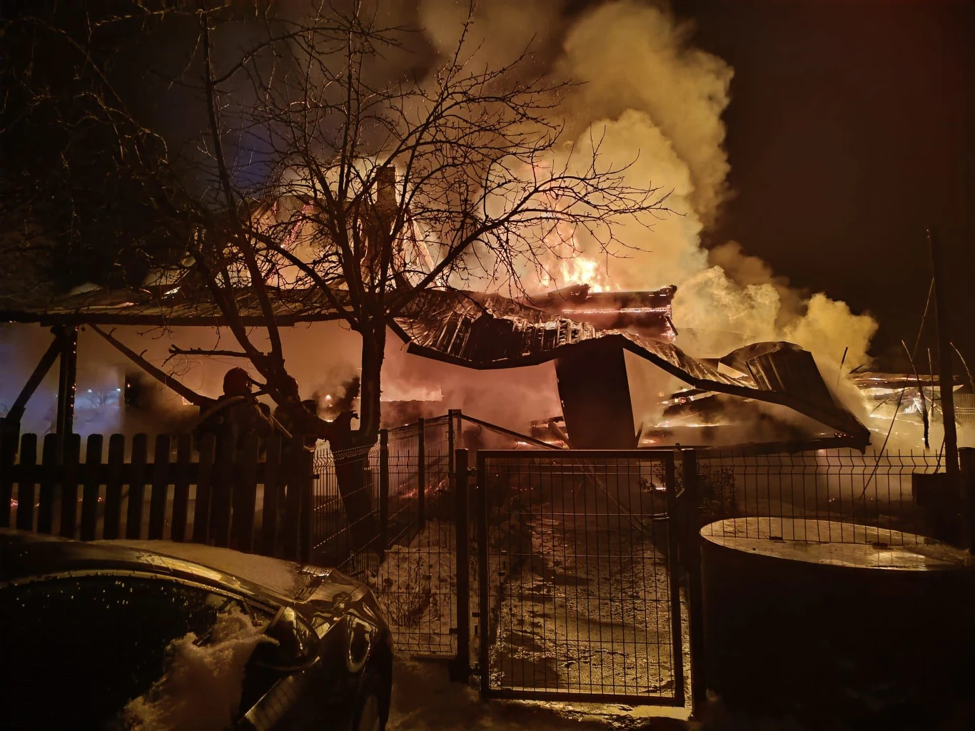 Tragiczny pożar domu na Podlasiu Duży pożar obejmujący budynek, płomienie i gęsty dym wydobywają się spod zawalającego się dachu, na pierwszym planie widoczny ogrodzenie oraz sylwetka osoby próbującej opanować sytuację.