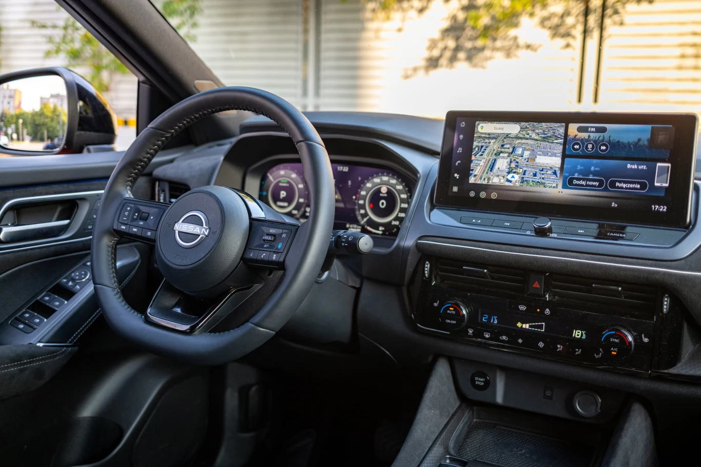 Nowoczesne wnętrze samochodu Nissan z cyfrowym wyświetlaczem zegarów, dużym ekranem dotykowym z nawigacją oraz systemem klimatyzacji dwustrefowej. Skórzana kierownica z wieloma przyciskami sterującymi oraz nowoczesna konsola środkowa podkreślają komfor...