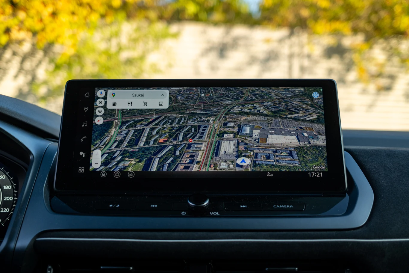 Kolorowy ekran w samochodzie wyświetlający mapę satelitarną miasta z nawigacją, dobrze widoczne szczegóły infrastruktury miejskiej, czas oraz pasek wyszukiwania.