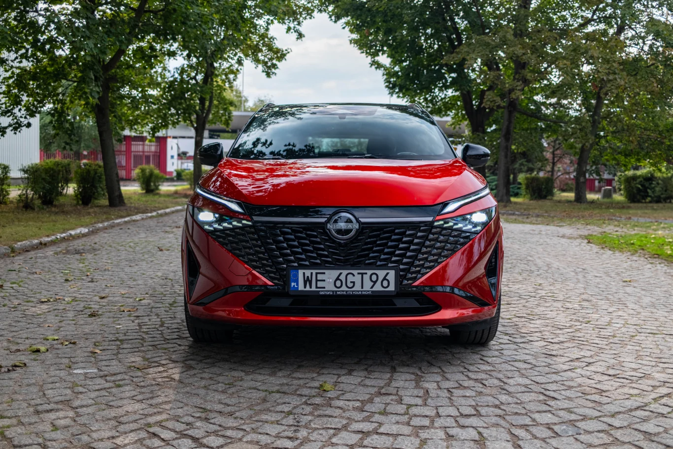 Czerwony samochód osobowy klasy kompaktowej stoi na brukowanej drodze, otoczony zielonymi drzewami, widoczny z przodu na tle miejskiego parku.