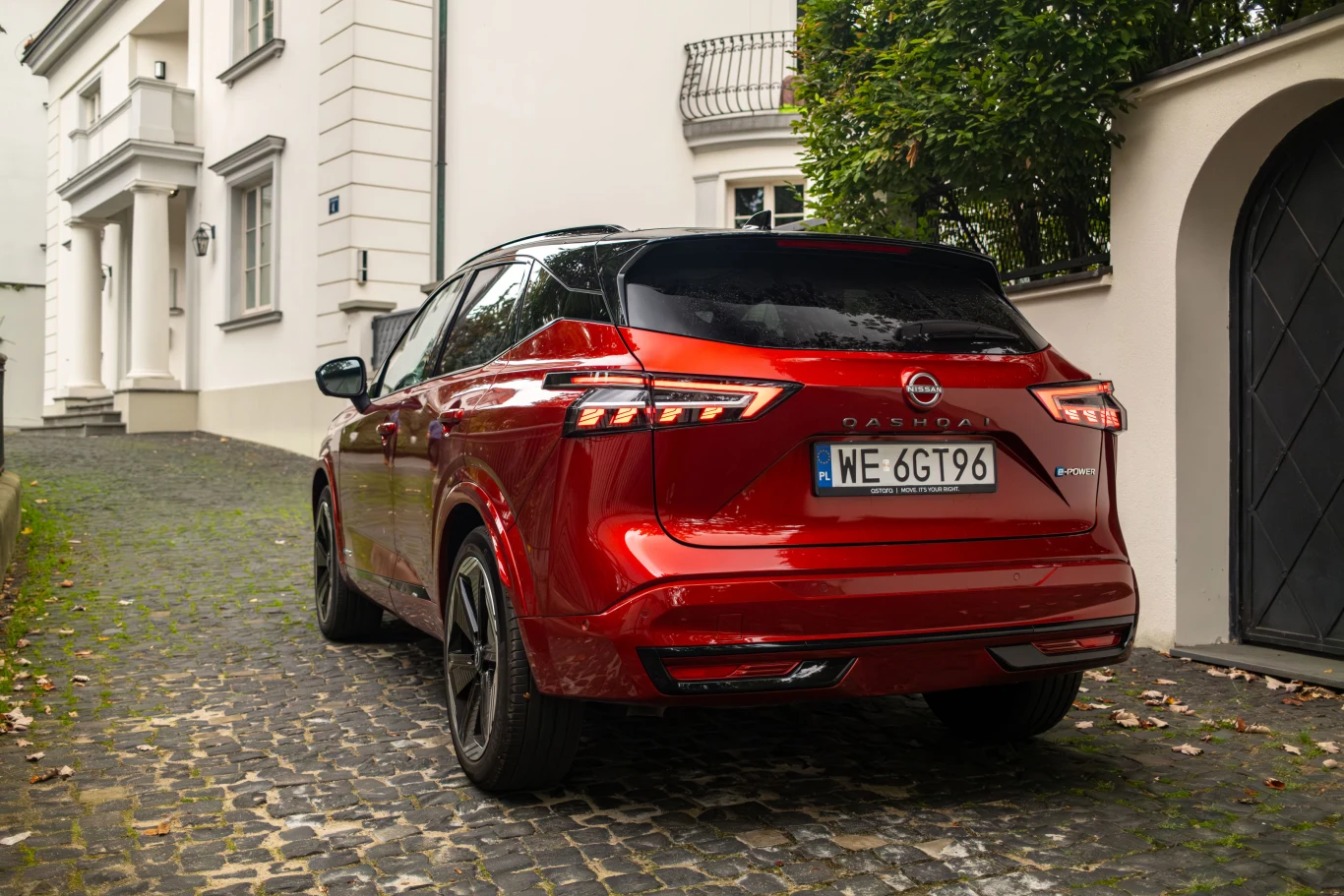Czerwony samochód Nissan Qashqai e-Power zaparkowany na brukowanej podjeździe przy eleganckiej białej willi, otoczony zielenią i klasyczną architekturą.