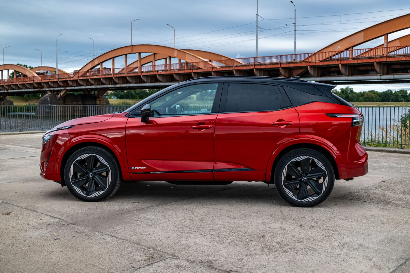 Czerwony nowoczesny SUV zaparkowany na tle mostu łukowego nad rzeką, z czarnymi elementami wykończenia i charakterystycznymi felgami, prezentowany z profilu na utwardzonym podłożu.