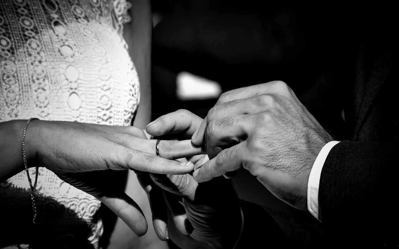 Mężczyzna zakłada kobiecie obrączkę na palec podczas ceremonii ślubnej, dłonie obojga są wyeksponowane na pierwszym planie, kobieta ma na sobie koronkową suknię.