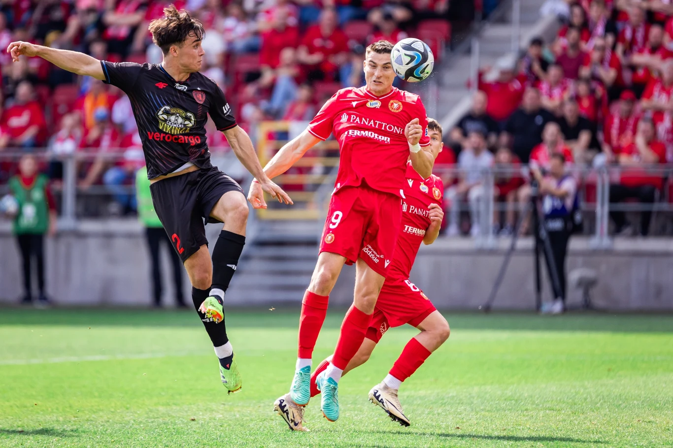 Dwóch piłkarzy rywalizuje o piłkę na boisku, jeden z nich skacze wysoko, drugi przygotowuje się do przyjęcia piłki głową, w tle widoczni kibice na trybunach.